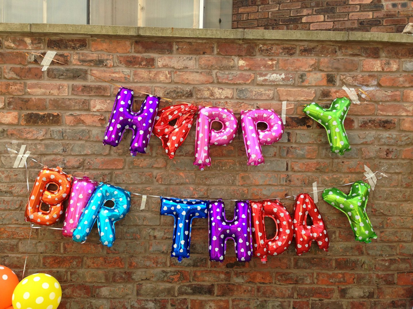 Multicolored foil balloons spell 'Happy Birthday' on a brick wall.